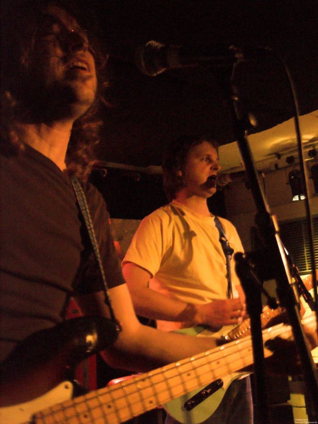 Eine Person in einem weißen Hemd singt in ein Mikrofon und spielt eine grüne Gitarre, während sie neben einer anderen Person in einem schwarzen Hemd und Brille steht, die ebenfalls singt und eine schwarze Gitarre spielt, mit Bühnenbeleuchtung und anderen Instrumenten im Hintergrund.