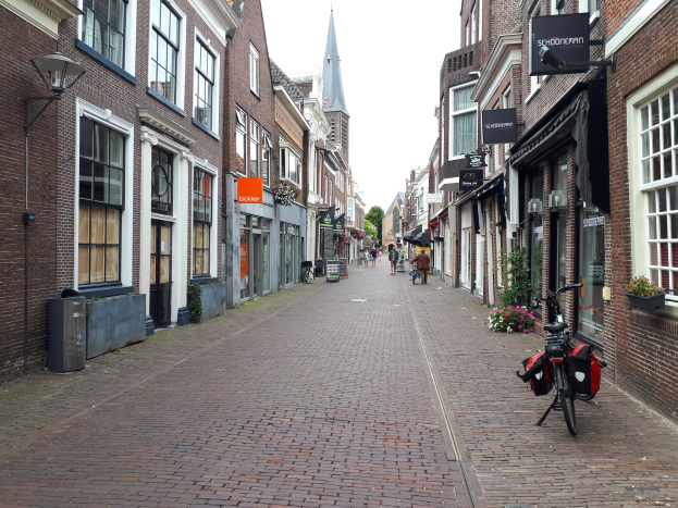 Ein Fahrrad, das auf einer Stadtstraße in Delft, Niederlande, geparkt ist und von Gebäuden mit Fenstern, Blumentöpfen, Schildern, einem Mülleimer, Bäumen und einem klaren blauen Himmel flankiert wird.