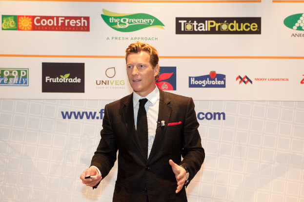 Ein Mann in einem schwarzen Blazer hält ein Objekt und spricht, mit einem weißen Banner und einer weißen Tafel mit organisationalen Logos im Hintergrund.