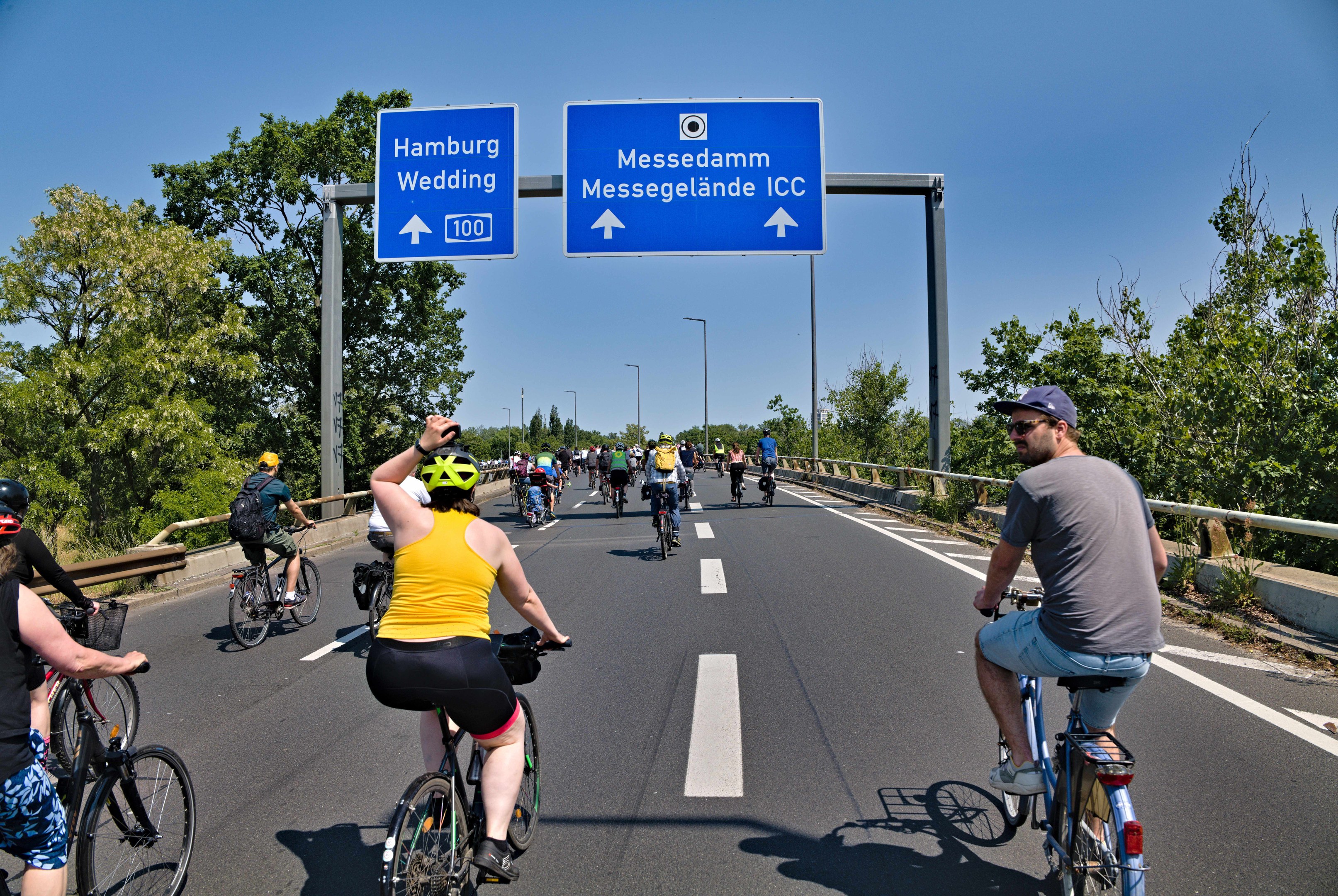 Gruppe von Radfahrern in Helmen auf einer Straße mit Bäumen auf einer Seite und einem Geländer auf der anderen, Laternen im Hintergrund unter einem klaren blauen Himmel, mit einem Schild oben, das eine Radtour in Hamburg anzeigt.