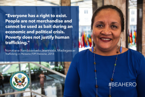 Eine Frau in einem blauen Kleid steht lachend vor einem Gebäude mit einer Aussage über Menschenhandel, umgeben von Menschen, Pflanzen und Flaggen.