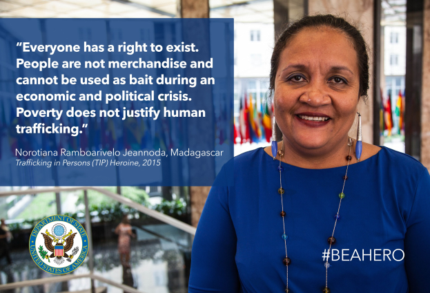 Eine Frau in einem blauen Kleid steht lachend vor einem Gebäude mit einer Aussage über Menschenhandel, umgeben von Menschen, Pflanzen und Flaggen.