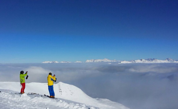 Zwei Personen stehen auf Snowboards in einer verschneiten Landschaft, mit Gegenständen in den Händen, mit Hügeln und Himmel im Hintergrund.