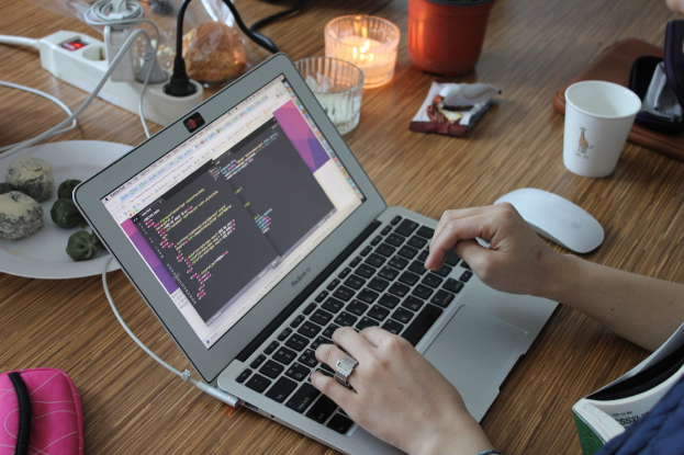 Ein Tisch mit einem Laptop, an dem eine Person tippt, eine Steckdose mit festen Kabeln, ein Teller mit Essen, eine Tasche, zwei Gläser, eine Glaskerze, eine Verpackung, ein Becher und eine Maus.