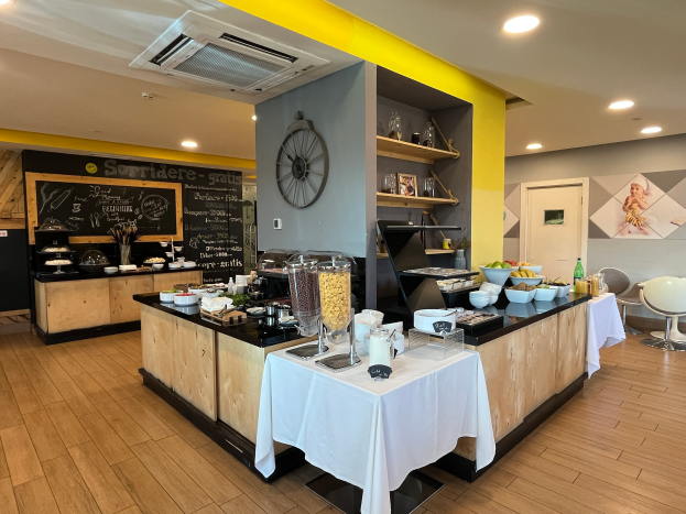 Ein Restaurantinnenraum mit Tischen und Stühlen, Speisen, einer Tafel mit Text, einer Wanduhr, Deckenleuchten, einer Klimaanlage und einer Tür im Hintergrund, befindet sich im Hôtel de Ville in Paris.