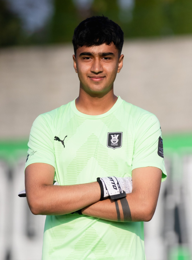 Ein junger Mann in einem grünen T-Shirt auf einem Fußballfeld mit verschränkten Armen und einem Lächeln, wahrscheinlich ein Torwart, mit einem unscharfen Hintergrund.