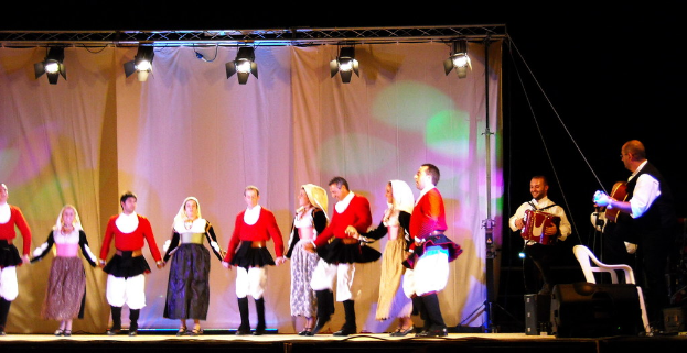 Eine Bühnenperformance mit Menschen auf der Bühne, einem Vorhang im Hintergrund, Lampen oben, Musikern rechts und einem weißen Stuhl unten rechts.