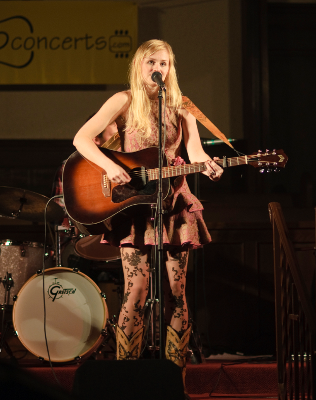 Eine Frau auf der Bühne singend und Gitarre spielend bei einem Konzert, mit einer Band im Hintergrund und einem Bildschirm an der Wand hinter ihr.