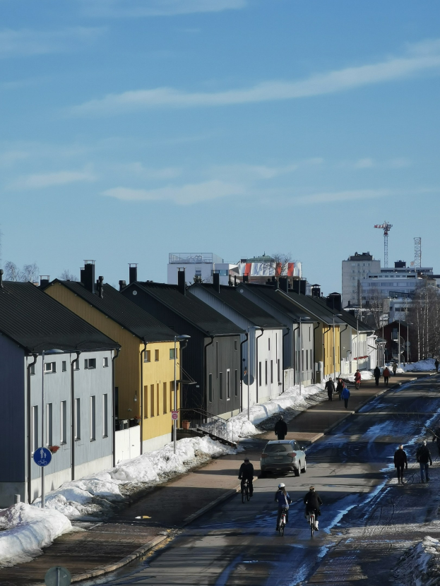 Eine Gruppe von Menschen, die auf Fahrrädern eine schneebedeckte Straße entlangfahren, gesäumt von Gebäuden, Bäumen, Pfählen und Schildern unter einem bewölkten Himmel.