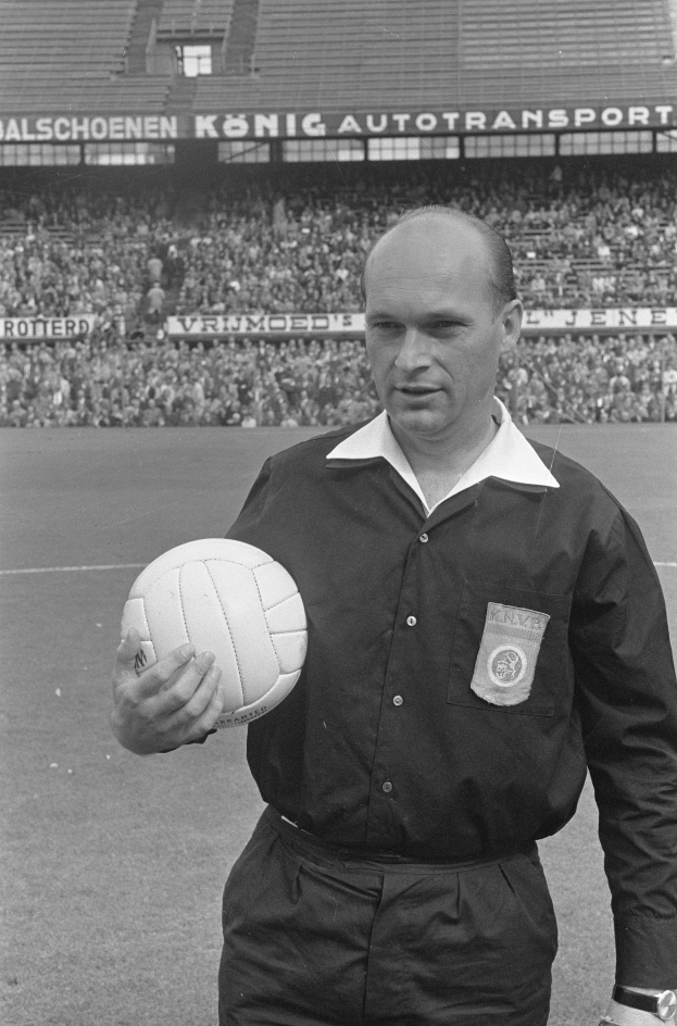 Ein Schwarz-Weiß-Foto eines Schiedsrichters auf einem Fußballfeld, der einen Ball hält, mit Zuschauern und Schildern mit Text im Hintergrund.