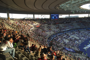 Große Menschenmenge in einem Stadion bei einem Fußballspiel mit einer Bühne, Fahnen und einem Bildschirm im Hintergrund, identifiziert als Allianz Arena in München, Deutschland.