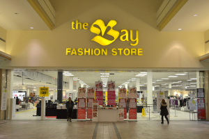Außenansicht eines Bay Fashion Stores in einem Einkaufszentrum mit Kunden, Wandtafeln, tragenden Säulen, Deckenbeleuchtung und diversen Hintergrund-Elementen.