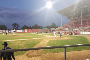 Baseballspiel im Gange mit Zuschauern auf den Stadionrängen, Bäume und Stadioninfrastruktur sind unter einem klaren blauen Himmel sichtbar.