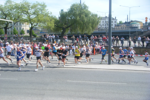 Eine Gruppe von Menschen, die bei einem Marathon auf einer Straße laufen, umgeben von einem Metallzaun, einem Tor, einer Schleife, die an einem Pfahl befestigt ist, Zuschauern auf dem Gehweg, einer Metallabsperrung, Pfählen, Straßenpfählen, Schildern, einer Brücke, Gebäuden mit Fenstern, Bäumen und einem bewölkten Himmel.