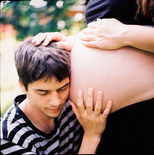 Ein Mann und eine Frau stehen nebeneinander, der Mann berüthrt zart den Bauch der Frau, mit einem unscharfen Hintergrund, der das Paar und ihr ungeborenes Kind betont.