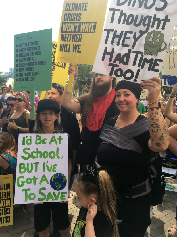 Eine Gruppe von Menschen mit Schildern, die vor einer Menge protestieren, mit Bäumen, Gebäuden und einem klaren blauen Himmel im Hintergrund.