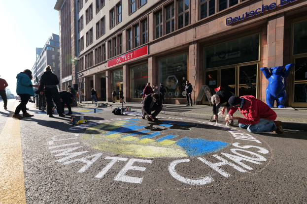 Menschen sitzen vor einem Gebäude mit Fenstern und Namensschildern während einer Klimawandel-Demonstration in Berlin, umgeben von Flaschen und anderen Gegenständen, mit Bäumen und einem klaren blauen Himmel im Hintergrund.
