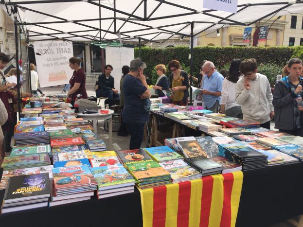 Eine Gruppe von Menschen steht um einen Tisch herum, der mit einem Tuch bedeckt ist und mit Büchern gefüllt ist, einige halten Bücher in der Hand, mit einem Banner mit Text, Pflanzen, Gebäuden mit Fenstern und einem klaren blauen Himmel im Hintergrund auf einer Buchmesse in Barcelona.