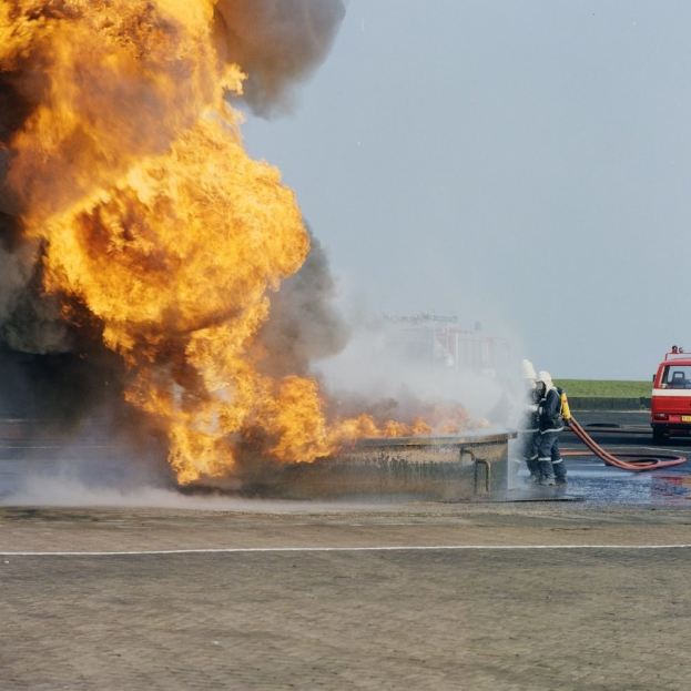 Ein Feuerwehrauto steht in Flammen am Rödchenrand, zwei Personen mit Helmen halten Schläuche in der Hand und ein Fahrzeug sowie der Himmel sind im Hintergrund zu sehen.
