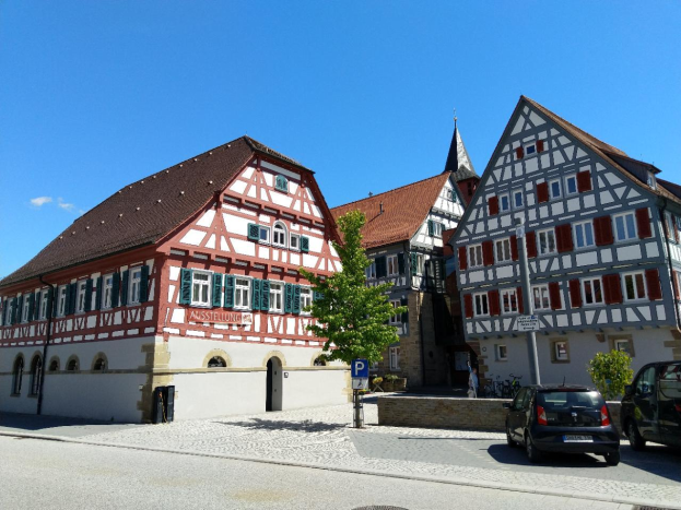 Eine Straße in Heidelberg, Deutschland, gesäumt von geparkten Autos, Gebäuden mit Fenstern, Bäumen und Schildern, unter einem Himmel voller weißer, flauschiger Wolken.