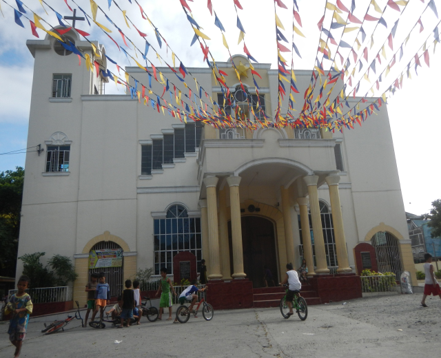 Eine Gruppe von Menschen, die vor einer weißen Kirche mit Fenstern, Säulen, Bögen und einem Kreuzsymbol Fahrräder fahren, umgeben von Pflanzen, einem Zaun, einer Fahne, Bäumen und einem bewölkten Himmel, mit bunten Fahnen, die an einem Seil befestigt sind.