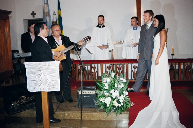 Eine HochzeitsSzene in einem Raum mit zwei Pfarrern, einem Brautpaar und drei Musikern; ein Podium mit Stoff davor, ein Tisch mit Kerzen dahinter, ein Kerzenhalter hinter dem Paar, ein Bukett, Lautsprecher, Taschen und roter Teppich auf dem Boden sowie Flaggen, eine Wand mit einem Kreuz, ein Schrank und ein Stuhl im Hintergrund.