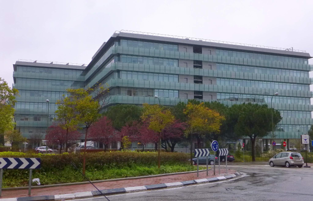 Großes modernes Gebäude mit zahlreichen Fenstern, als Europäische Zentralbank-Hauptquartier beschriftet, umgeben von Grünflächen, Straßeninfrastruktur und Fahrzeugen unter einem bewölkten Himmel.
