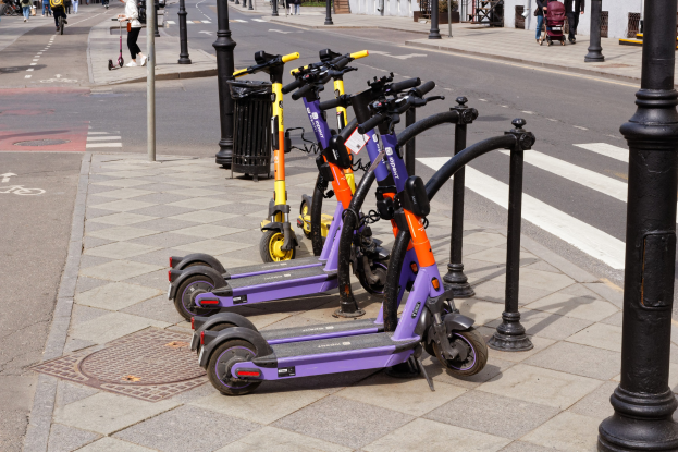 Eine Reihe von Elektro-Scootern, die an der Straßenseite geparkt sind, mit Menschen, die auf dem Gehweg laufen und Fahrräder fahren, einem Mülleimer im Vordergrund und Gebäuden, Bäumen und Pflanzen im Hintergrund.
