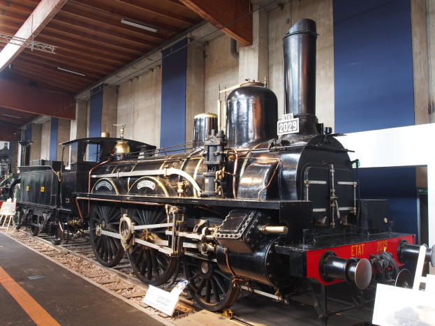 Eine große, alte Dampflokomotive in einem Museums Setting umgeben von Eisenbahnschienen, Informationsschildern und anderen Ausstellungsstücken, mit einer beleuchteten Decke und Wand im Hintergrund.