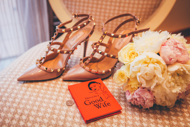 Ein Tisch zeigt ein Buch mit dem Titel "Wie man eine gute Ehefrau ist", einen Blumenstrauß und ein Paar Valentino-Schuhe, vor einem Vorhang.