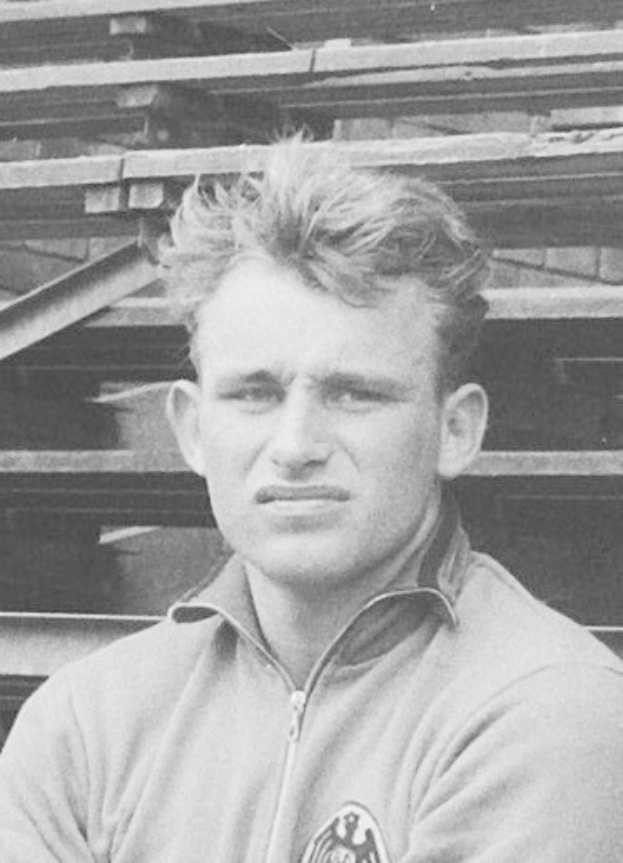 Schwarzes und weißes Foto eines Mannes in einer Jacke, der vor einem Haufen Holzplanken steht.