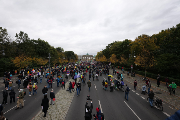 Eine große Gruppe von Menschen marschiert auf einer von Bäumen gesäumten Straße mit Laternenmasten, einige halten Kameras, während einer Protestdemo in Berlin, mit einem Gebäude und einem klaren Himmel im Hintergrund.