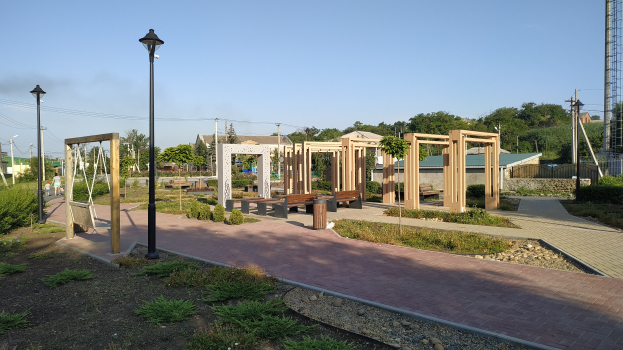 Parkszene mit einem Holzpfosten gesäumten Weg, Bänken, Pflanzen, Gras, Steinen, Straßenlaternen, Strommasten mit Drähten, Bäumen, Gebäuden und einem Turm im Hintergrund unter einem klaren blauen Himmel.