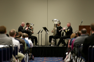 Eine Gruppe von Männern, die auf einer Bühne Blasinstrumente spielen, mit Notenständern, einem Podium mit Mikrofon und einem Publikum, das auf Stühlen sitzt, mit einer Wand im Hintergrund.