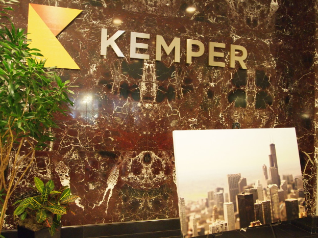 Eine Wand mit dem Wort "Kemper" darauf, flankiert von Pflanzen, mit einem Monitorbildschirm davor, der Gebäude anzeigt.