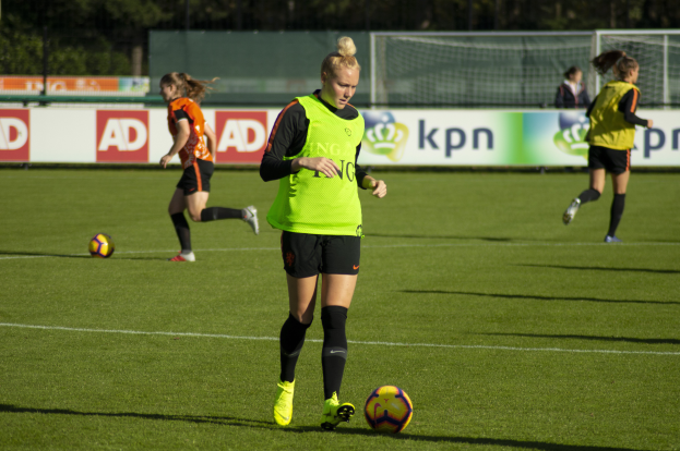 Eine Gruppe von Frauen in Fußballtrikots, die auf einem von Bäumen umgebenen Feld spielen, mit Bannern im Hintergrund und einem Netz im Vordergrund.