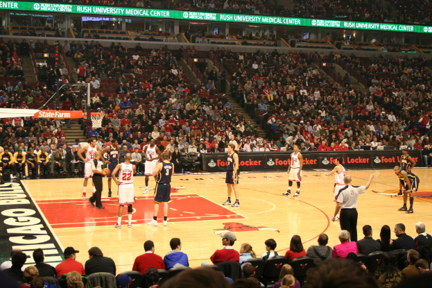 Eine Gruppe von Menschen in einer Arena, einige halten einen Basketball, andere schauen von den Rängen und Werbetafeln sind im Hintergrund zu sehen.