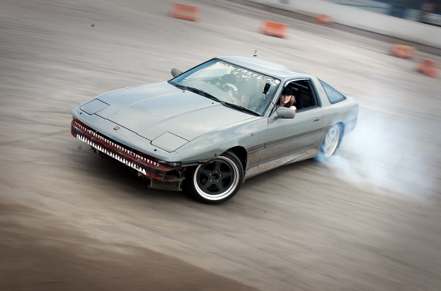 Eine Person mit Helm fährt ein Auto auf einer Rennstrecke, wobei Rauch aufsteigt, umgeben von orangefarbenen Objekten mit einem unscharfen Hintergrund, der hohe Geschwindigkeit andeutet.