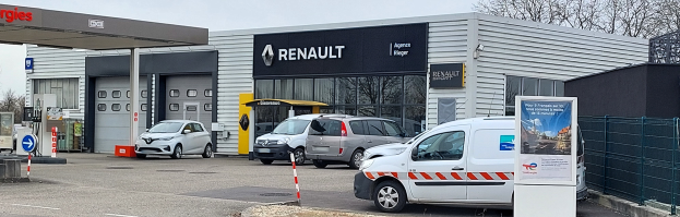 Ein Renault-Autohaus mit parkenden Autos vorne, umgeben von einem Metallzaun, einem Schild, Pfählen, einer Texttafel, Bäumen und einem bewölkten Himmel.