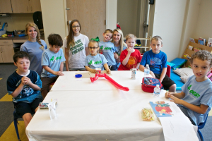 Eine Gruppe von Kindern und drei Frauen, einige lächeln, mit Schränken, einer Wand, einer Tür und Kissen im Hintergrund.