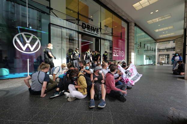 Eine Gruppe von Menschen sitzt vor einer Volkswagen-Verkaufsstelle mit Glaswänden auf dem Boden und hält ein Transparent.