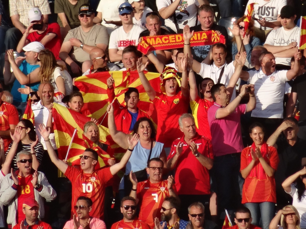 Eine Gruppe von Menschen in einem Stadion, die ihre Hände heben, einige tragen Kappen und Schutzbrillen, halten Fahnen und Schilder mit Text, im Hintergrund sind Geländer zu sehen.
