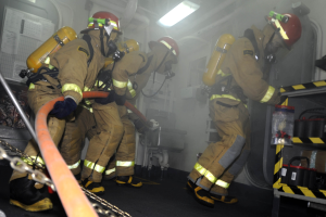 Feuerwehrleute in Schutzbekleidung und Helmen arbeiten an einem Feuerlöschfahrzeug, mit Ausrüstungsgestellen und Hintergrundinfrastruktur im Hintergrund.