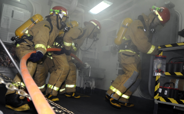 Feuerwehrleute in Schutzbekleidung und Helmen arbeiten an einem Feuerlöschfahrzeug, mit Ausrüstungsgestellen und Hintergrundinfrastruktur im Hintergrund.