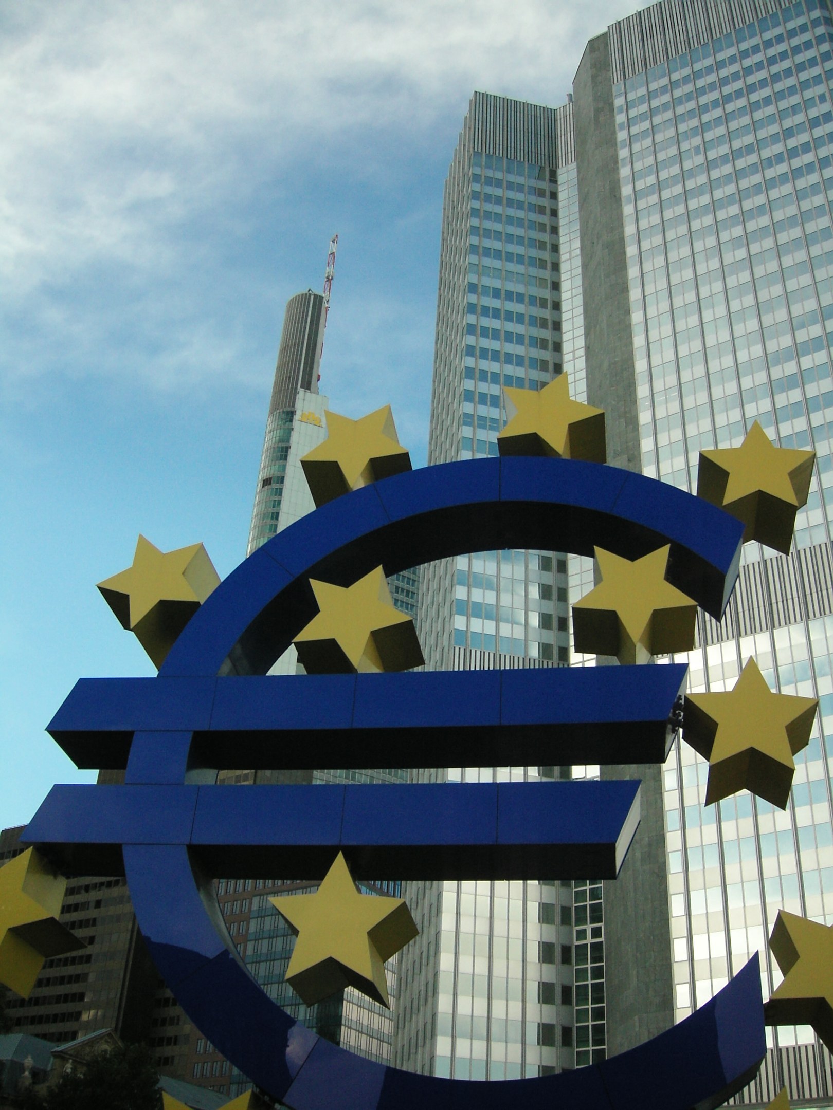Eurozeichen vor dem Gebäude der Europäischen Zentralbank in Frankfurt, Deutschland, mit Bäumen und einem bewölkten Himmel im Hintergrund.