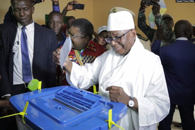 Ein Mann in einem weißen Anzug und Hut legt seine Stimme in eine blaue Wahlurne, während er von Menschen mit Kameras und Handys umgeben ist.