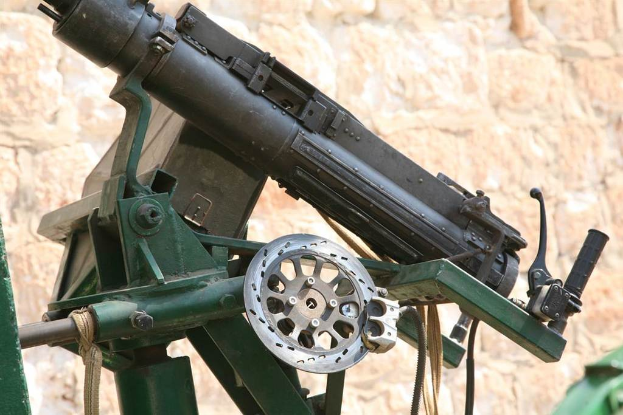 Eine Maschinengewehr auf einem Stahlgestell mit einem Griff auf der rechten Seite, Seilen und Kabeln unten und einer Wand im Hintergrund.