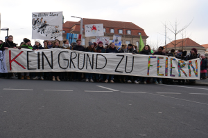 Demonstranten mit einem Transparent "Kein Grund zur Feier" gegen deutsche Sparmaßnahmen, mit Straßeninfrastruktur und Gebäuden im Hintergrund.