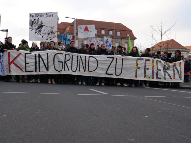Demonstranten mit einem Transparent "Kein Grund zur Feier" gegen deutsche Sparmaßnahmen, mit Straßeninfrastruktur und Gebäuden im Hintergrund.