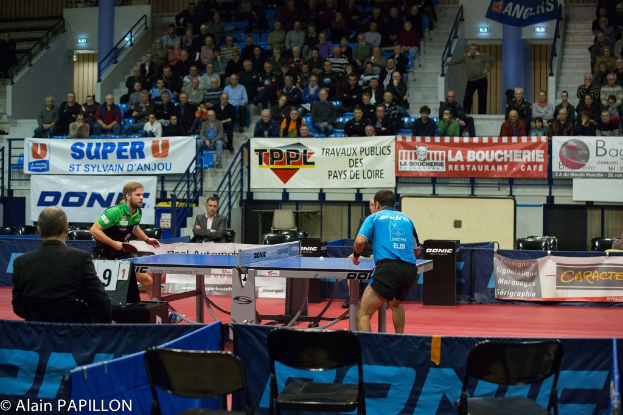 Zwei Männer spielen Tischtennis in einer Halle, umgeben von mehreren Zuschauern, einigen leeren Stühlen und Werbeplakaten.